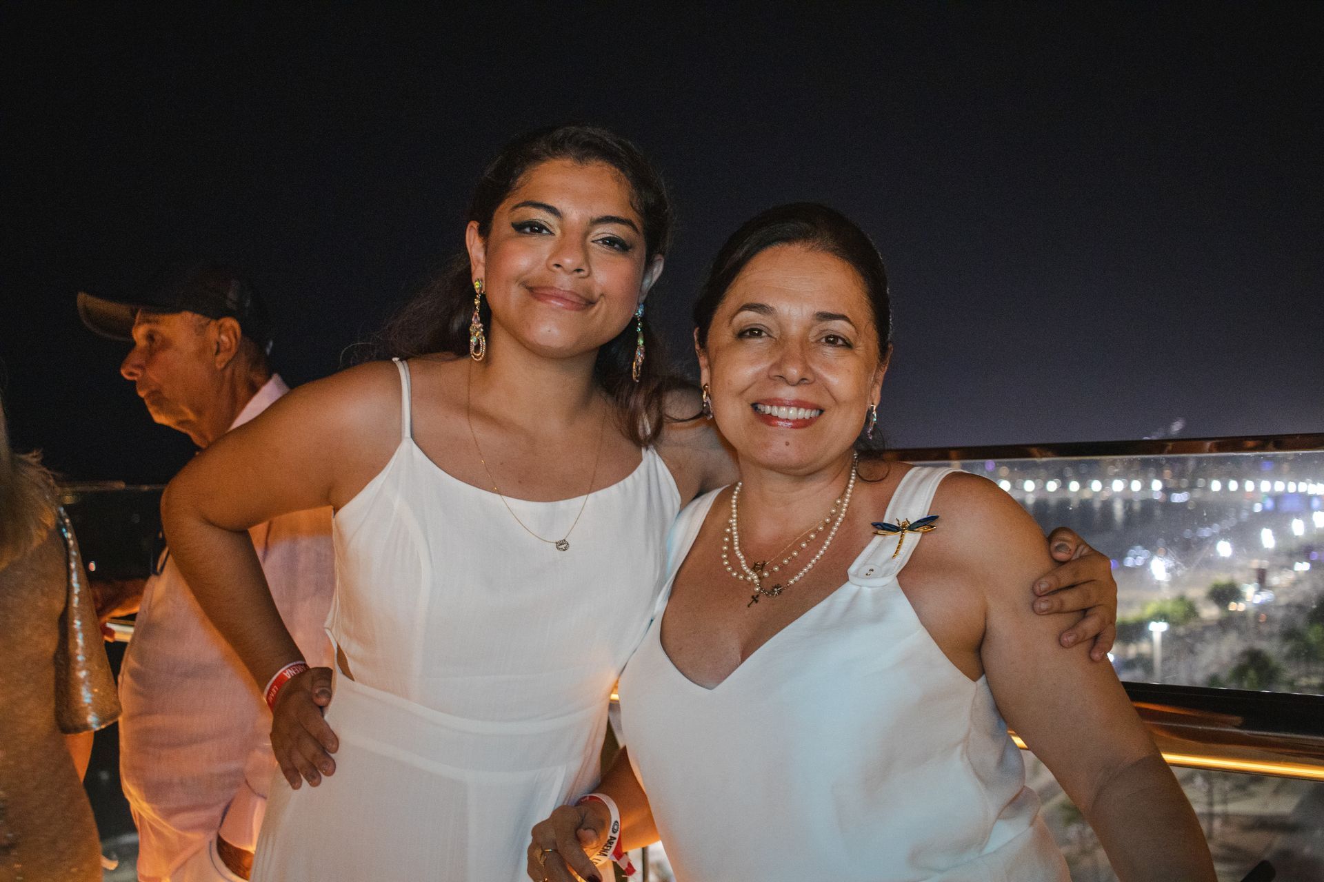 Duas pessoas sorrindo para uma foto contra um cenário noturno iluminado da cidade, ambas vestindo roupas brancas.