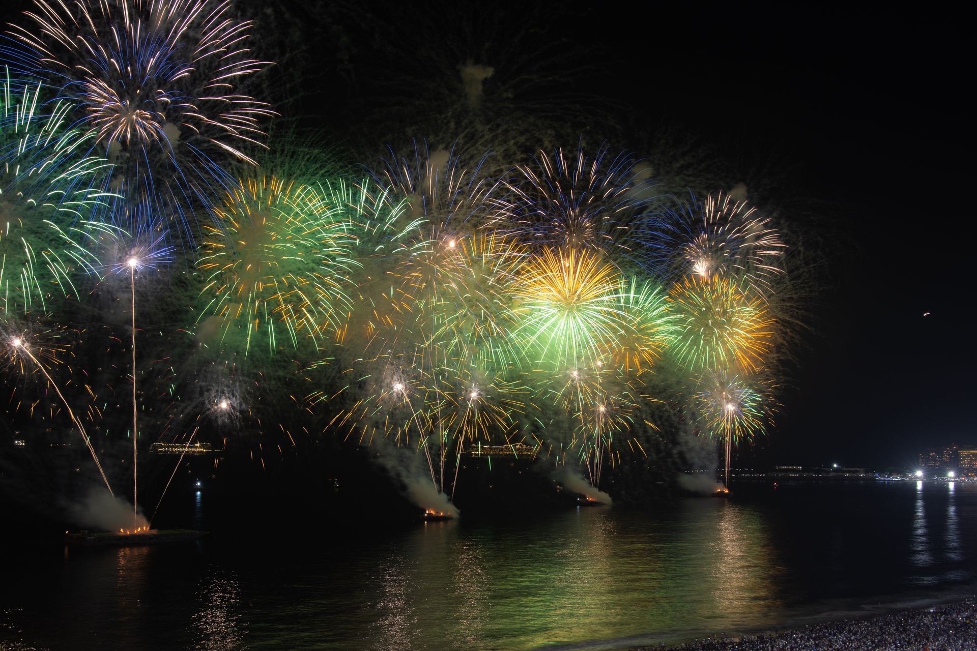 Fogos de artifício em tons vibrantes de verde e dourado explodem sobre a água escura, refletindo as luzes contra o céu noturno.