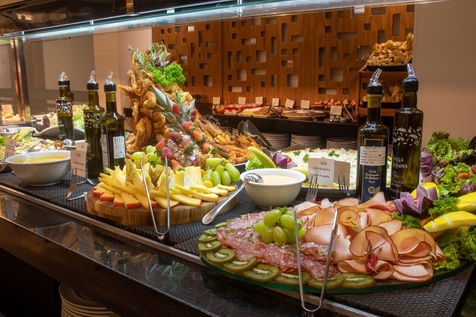 Um buffet com fatias de queijo, carnes curadas, uvas e diversas garrafas de azeite sobre um balcão escuro.