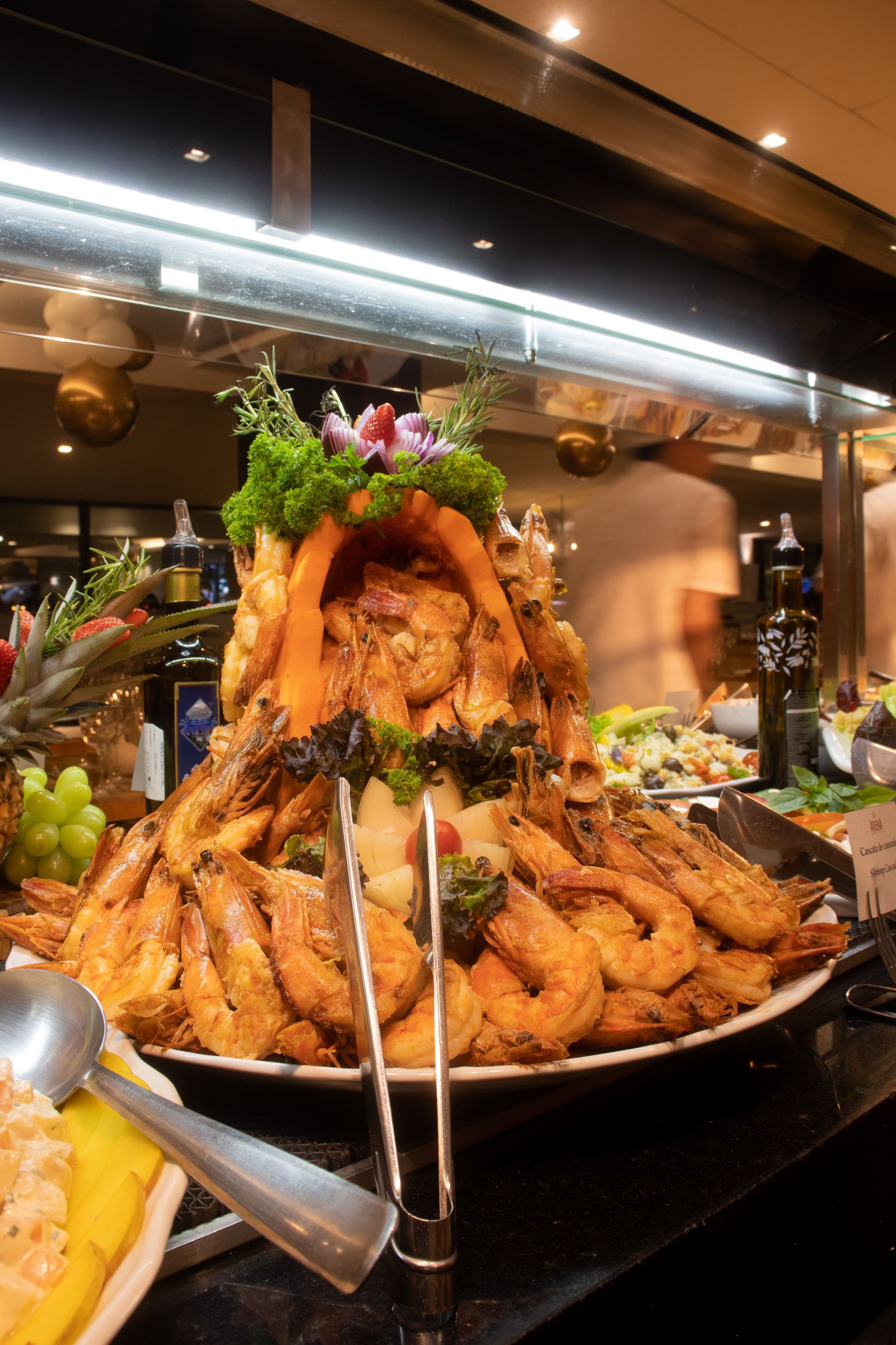 Uma grande e impressionante porção de camarões cozidos servida em uma travessa em um bufê, guarnecida com verduras e frutas.