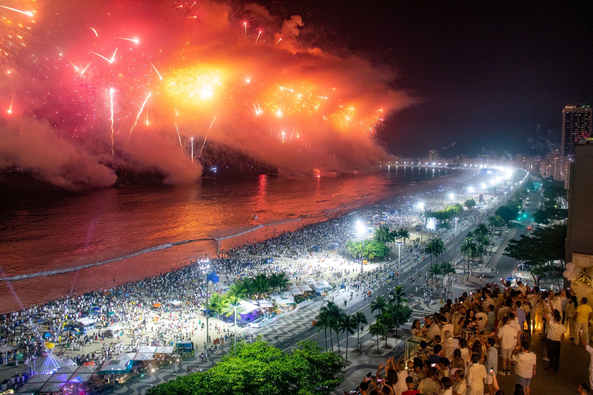 Fogos de artifício iluminam o céu noturno sobre uma praia lotada de Copacabana, com pessoas reunidas ao longo da costa e do calçadão.