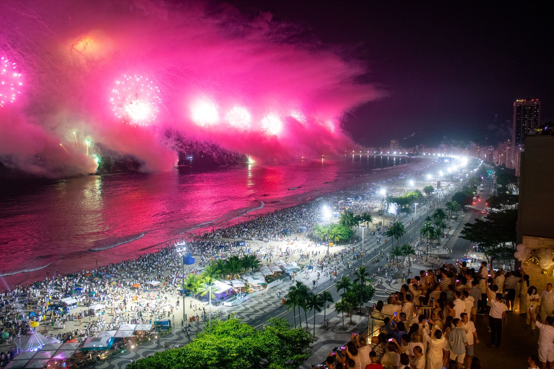 Uma praia à noite repleta de pessoas assistindo a vibrantes fogos de artifício rosa sobre o oceano e o horizonte da cidade.