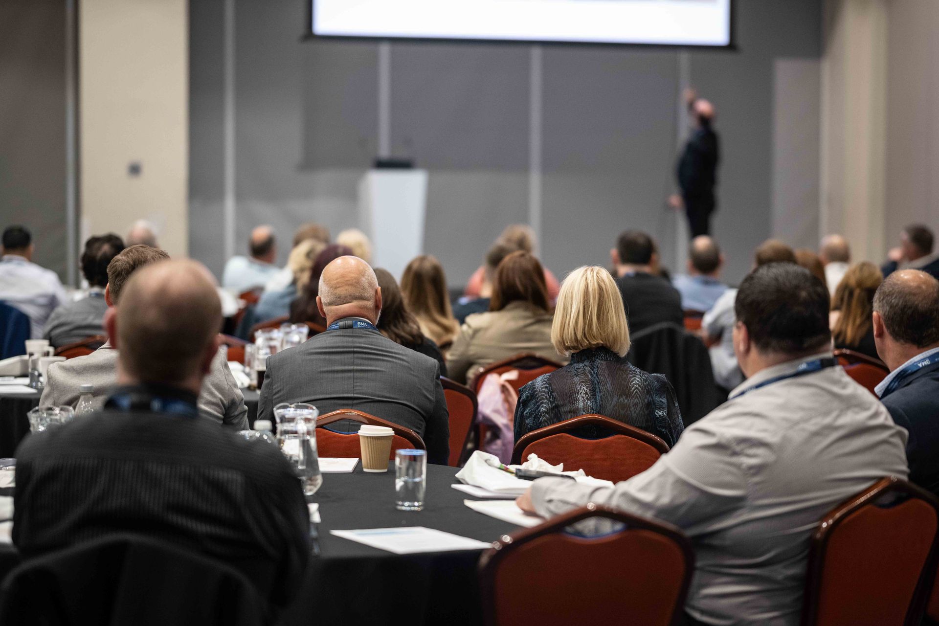 A group of people are sitting at tables in a conference room watching a presentation.