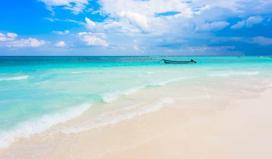 Playa de arena blanca con agua turquesa y un cielo azul con nubes; se ve una pequeña embarcación.