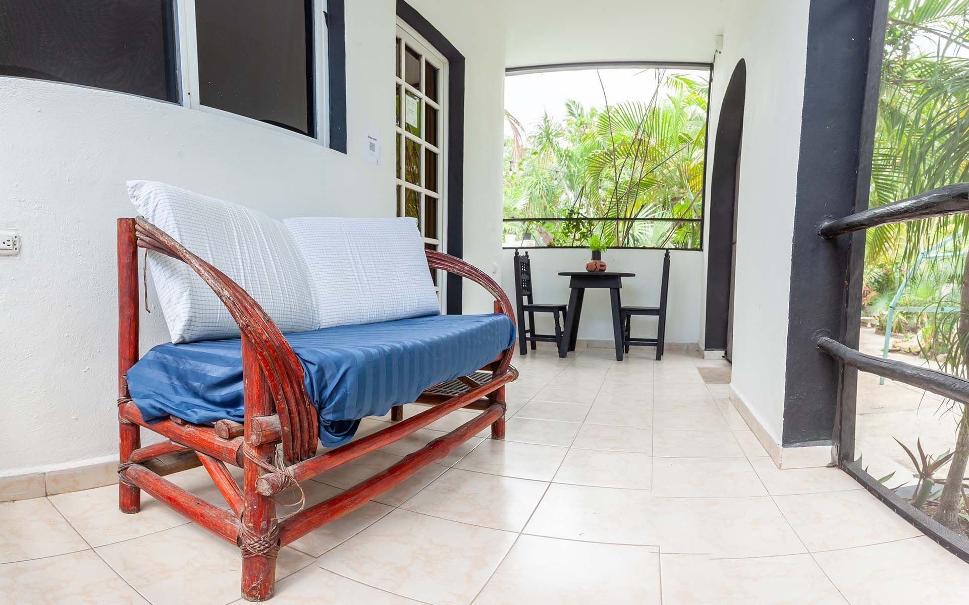 Patio cubierto con un sofá de madera roja, mesa y sillas; plantas al fondo.