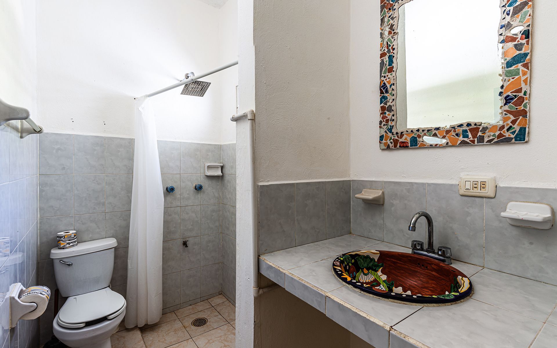 Baño con inodoro, ducha y lavabo con espejo de mosaico decorativo.