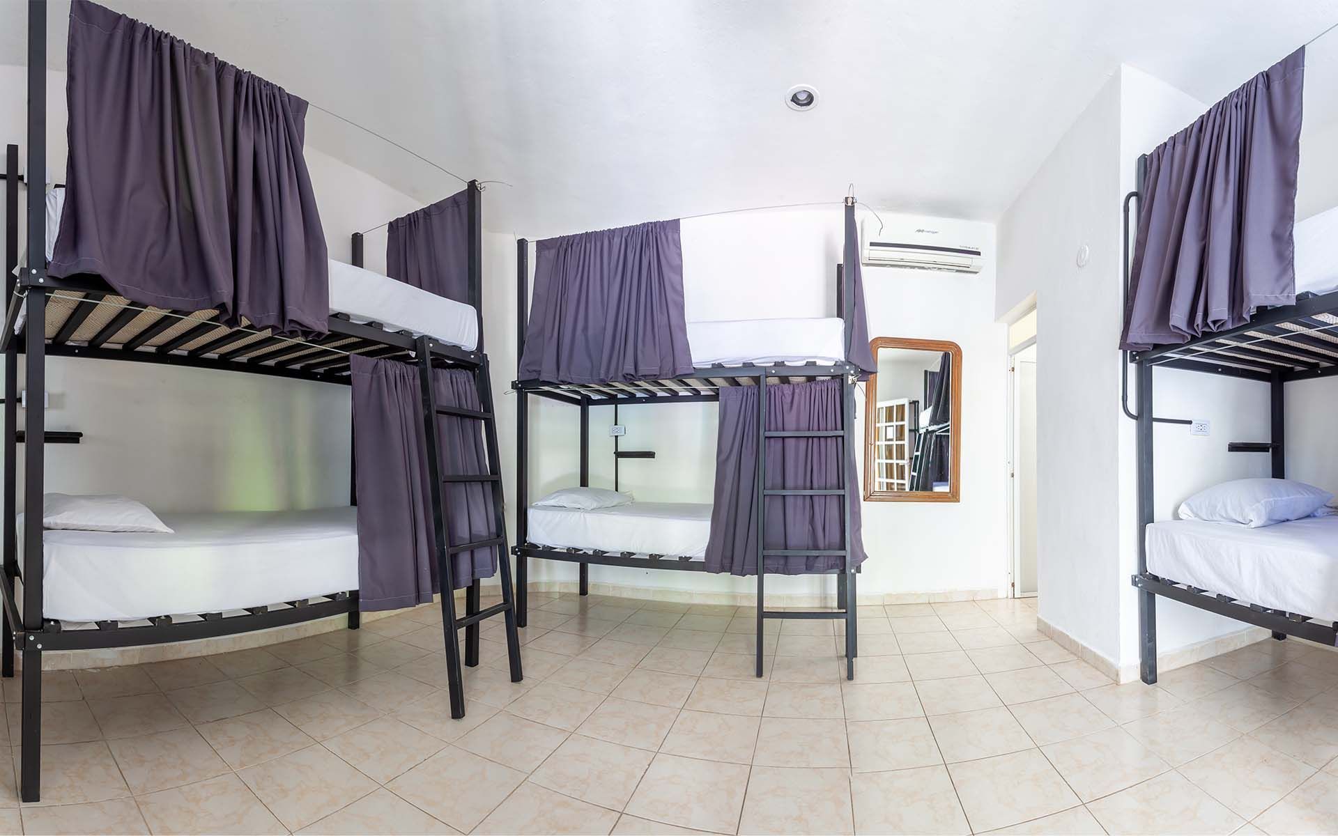 Habitación de albergue con varias literas negras, ropa de cama blanca y cortinas moradas.