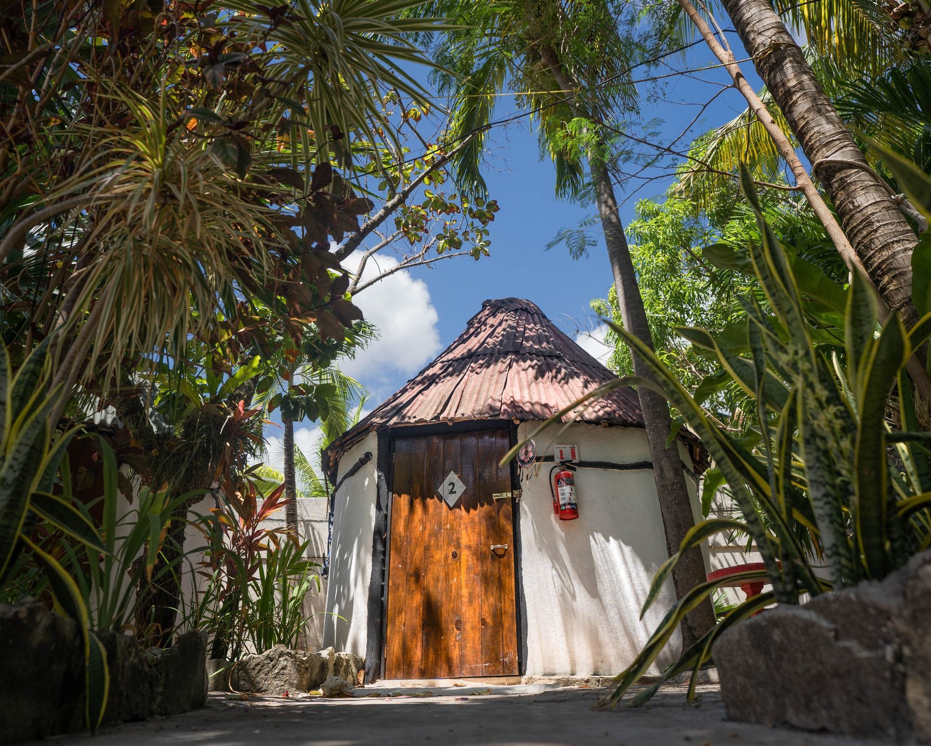 Pequeña cabaña con puerta de madera y techo de paja rodeada de exuberante follaje verde bajo un cielo azul.