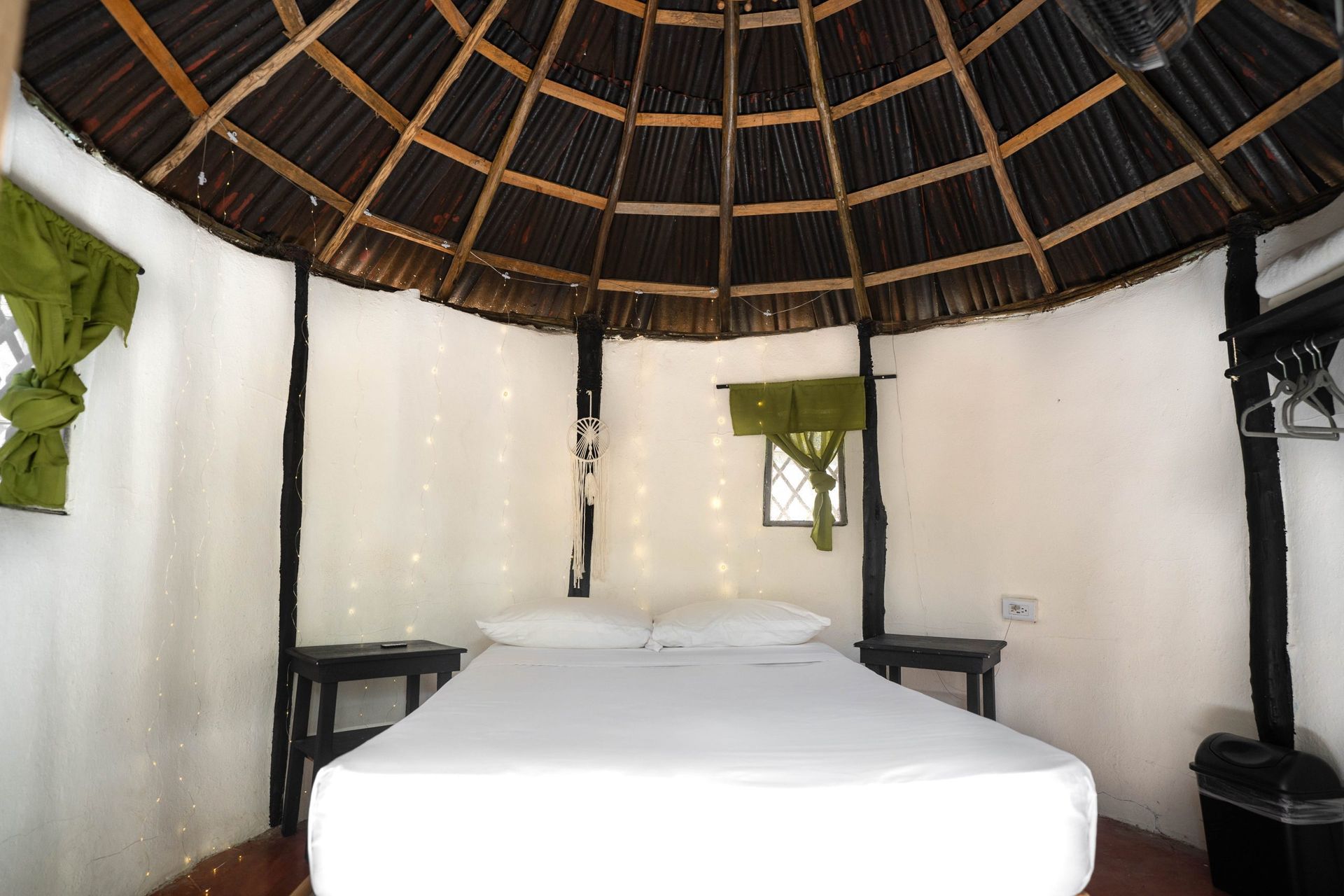 Dormitorio con techo redondo de paja. Paredes blancas, cama y mesitas de noche. Cortinas verdes.