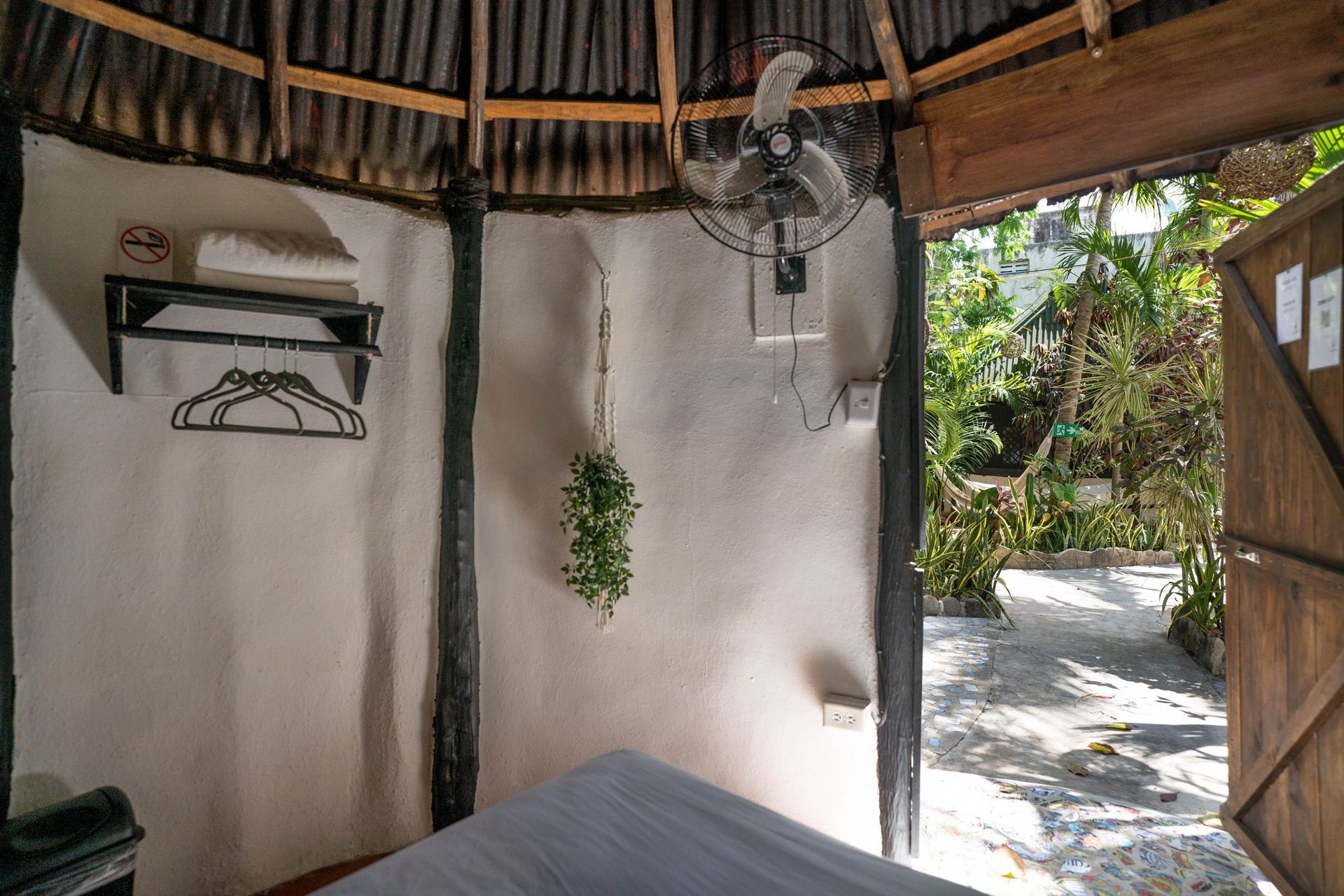 Vista interior de una pequeña habitación al aire libre; cama, ventilador, plantas colgantes, puerta de madera.