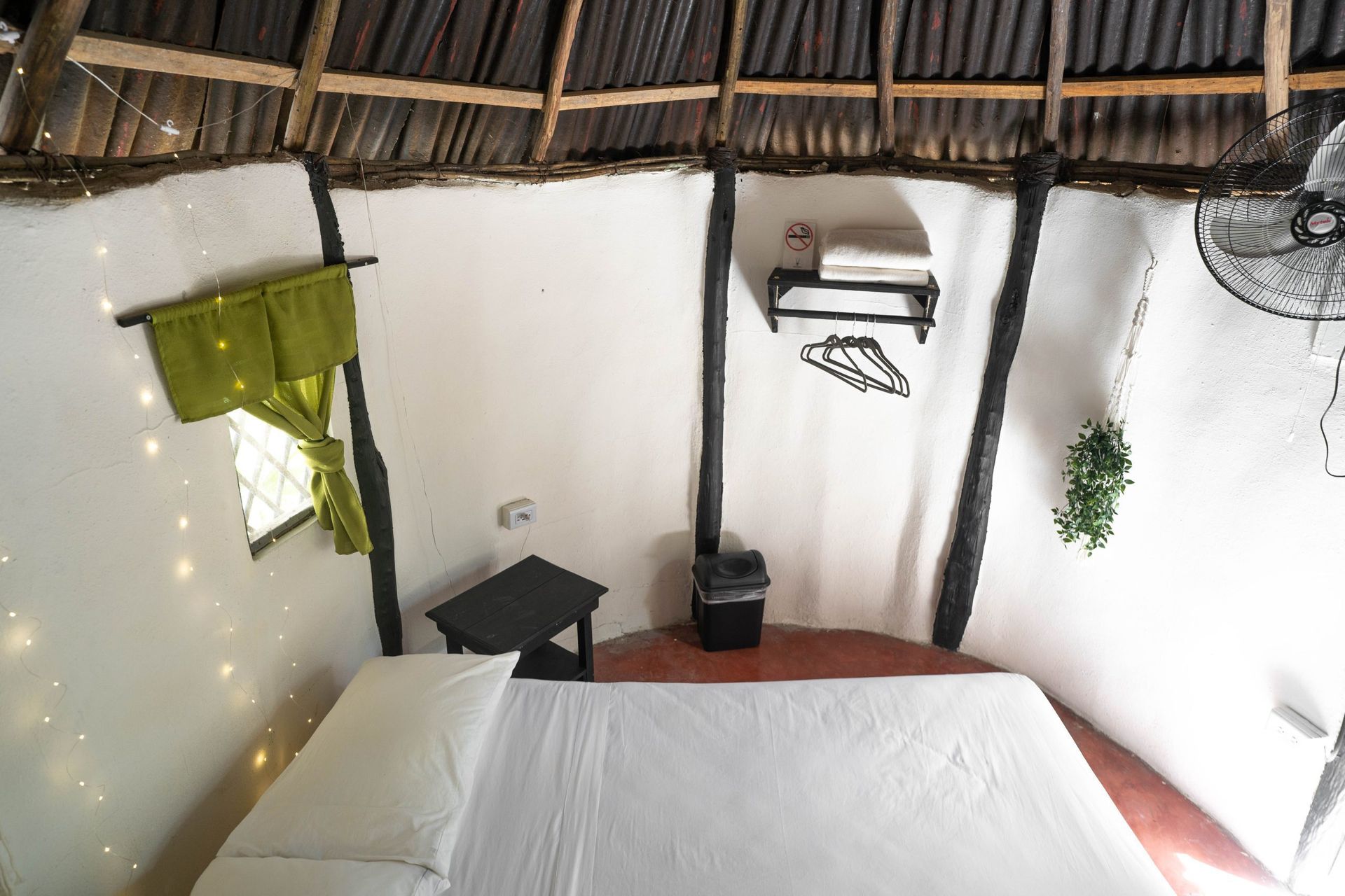 Interior de dormitorio con cama, estanterías, ventilador y cortinas verdes. Paredes blancas, vigas de madera y techo de paja.