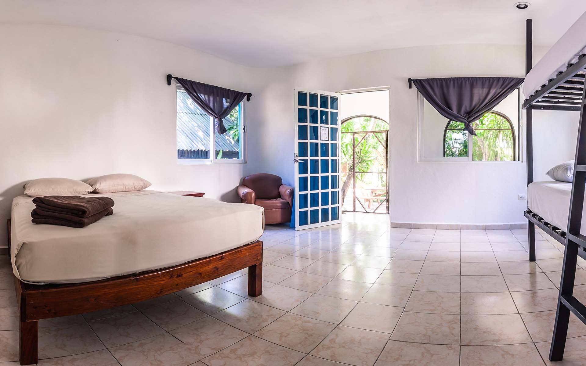 Habitación luminosa con cama doble, literas y puerta abierta a zona verde; suelo de baldosas, paredes blancas.