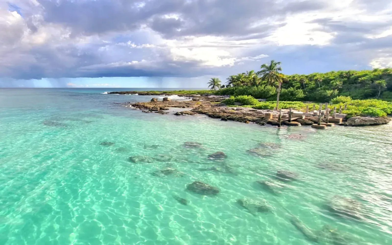 Océano turquesa con rocas y una costa verde bordeada de árboles bajo un cielo nublado.