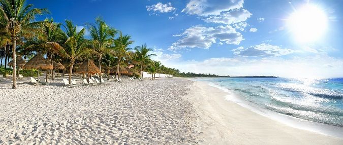 Una playa de arena blanca con palmeras y cabañas bajo un cielo azul brillante y sol.