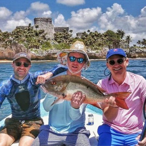 Tres hombres en un barco, sosteniendo un gran pez rojo cerca de ruinas antiguas.