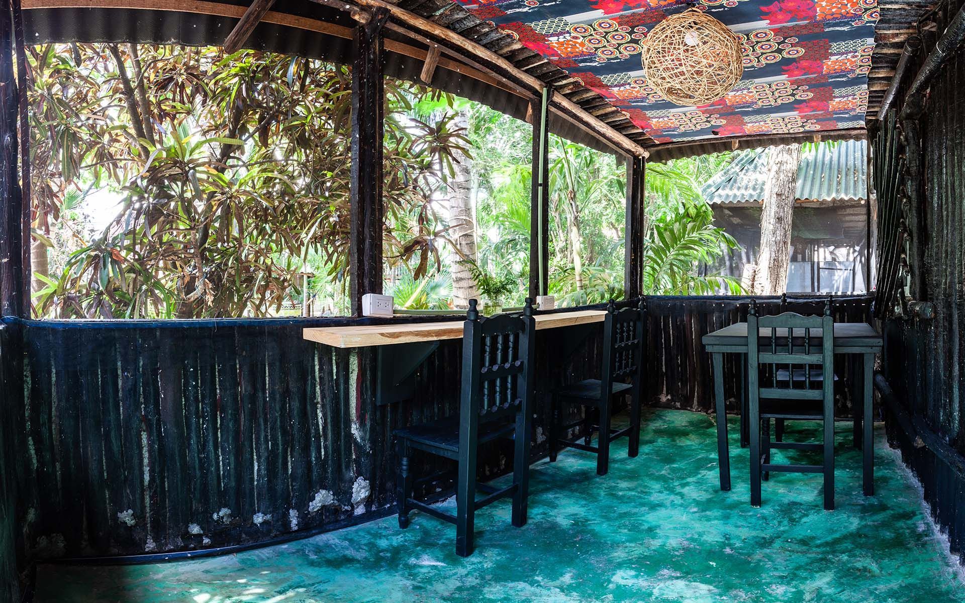 Comedor al aire libre con mesas y sillas negras, suelo verde y árboles visibles a través de las ventanas.