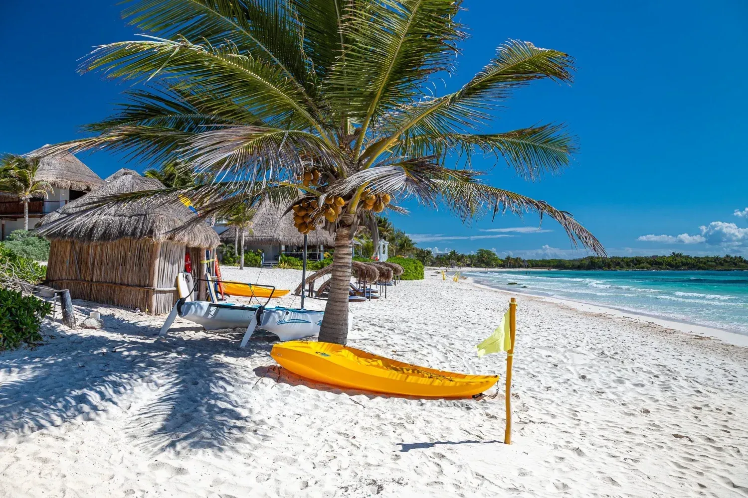 Escena de playa con kayak amarillo, palmeras y chozas de paja sobre arena blanca bajo un cielo azul.
