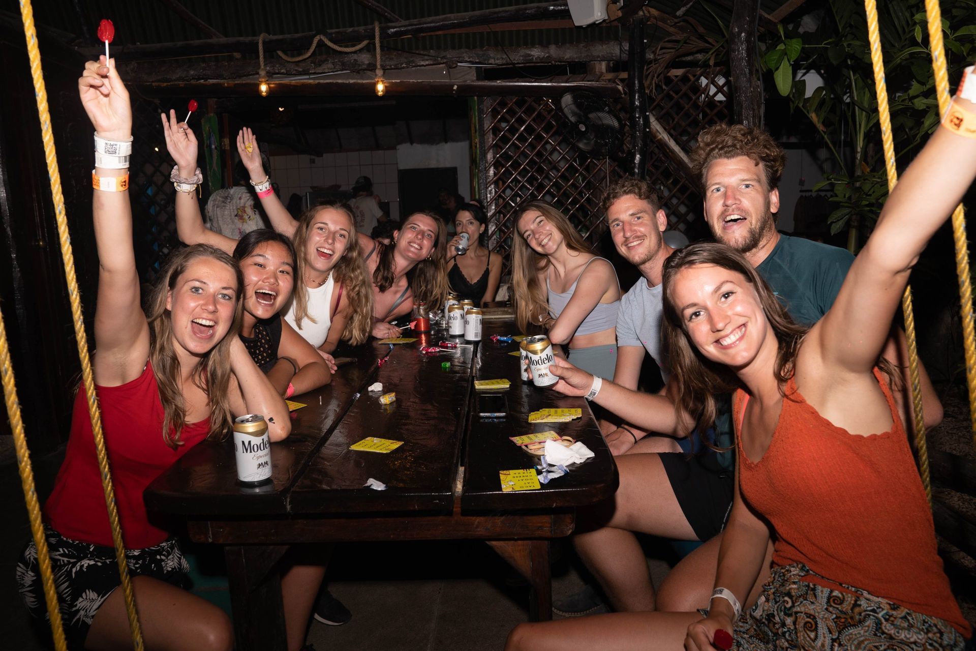 Grupo de personas en una mesa, sonriendo y levantando los brazos en un ambiente de fiesta; algunos sosteniendo bebidas.