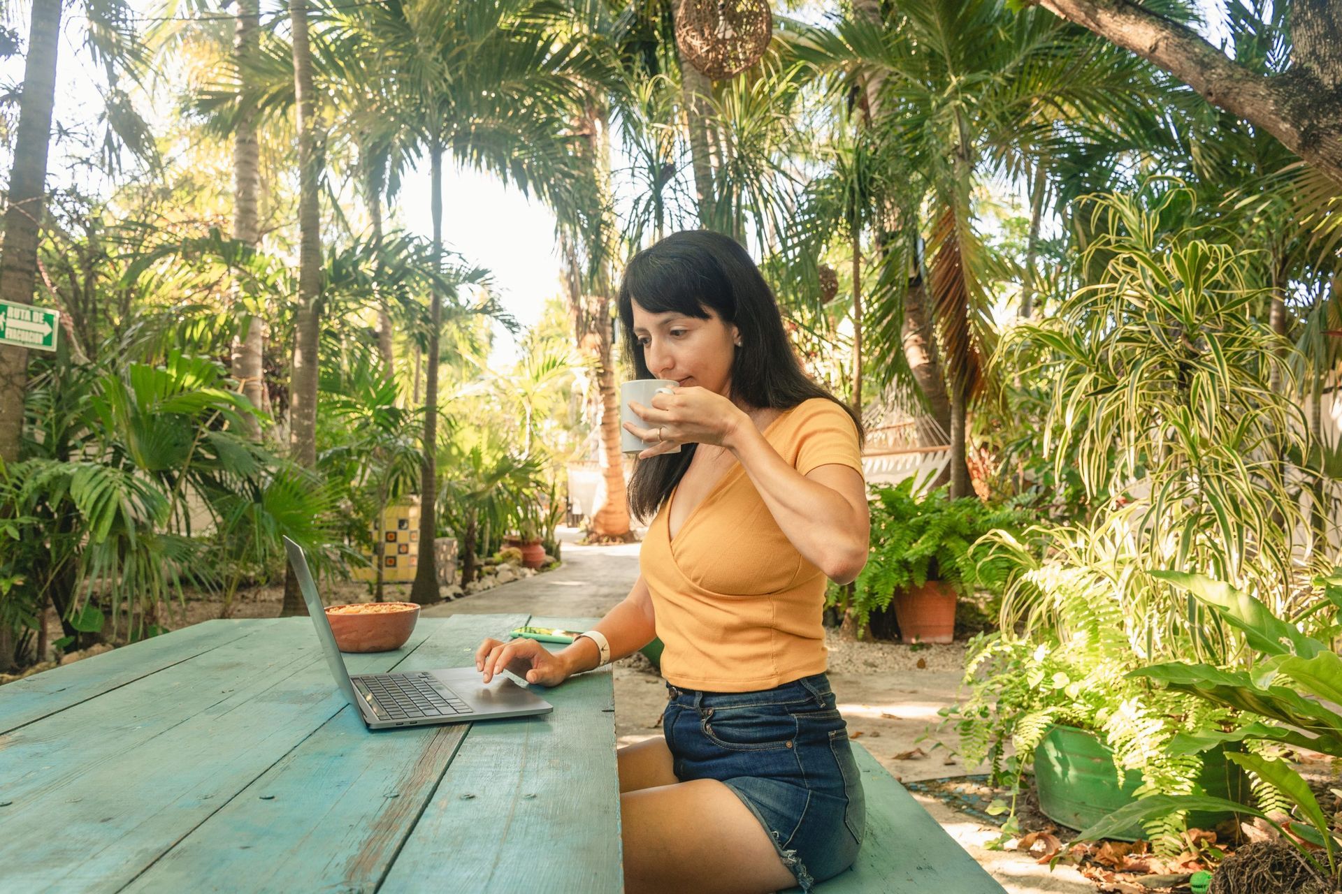 Mujer trabajando con una laptop al aire libre, bebiendo de una taza. Rodeada de exuberante vegetación.
