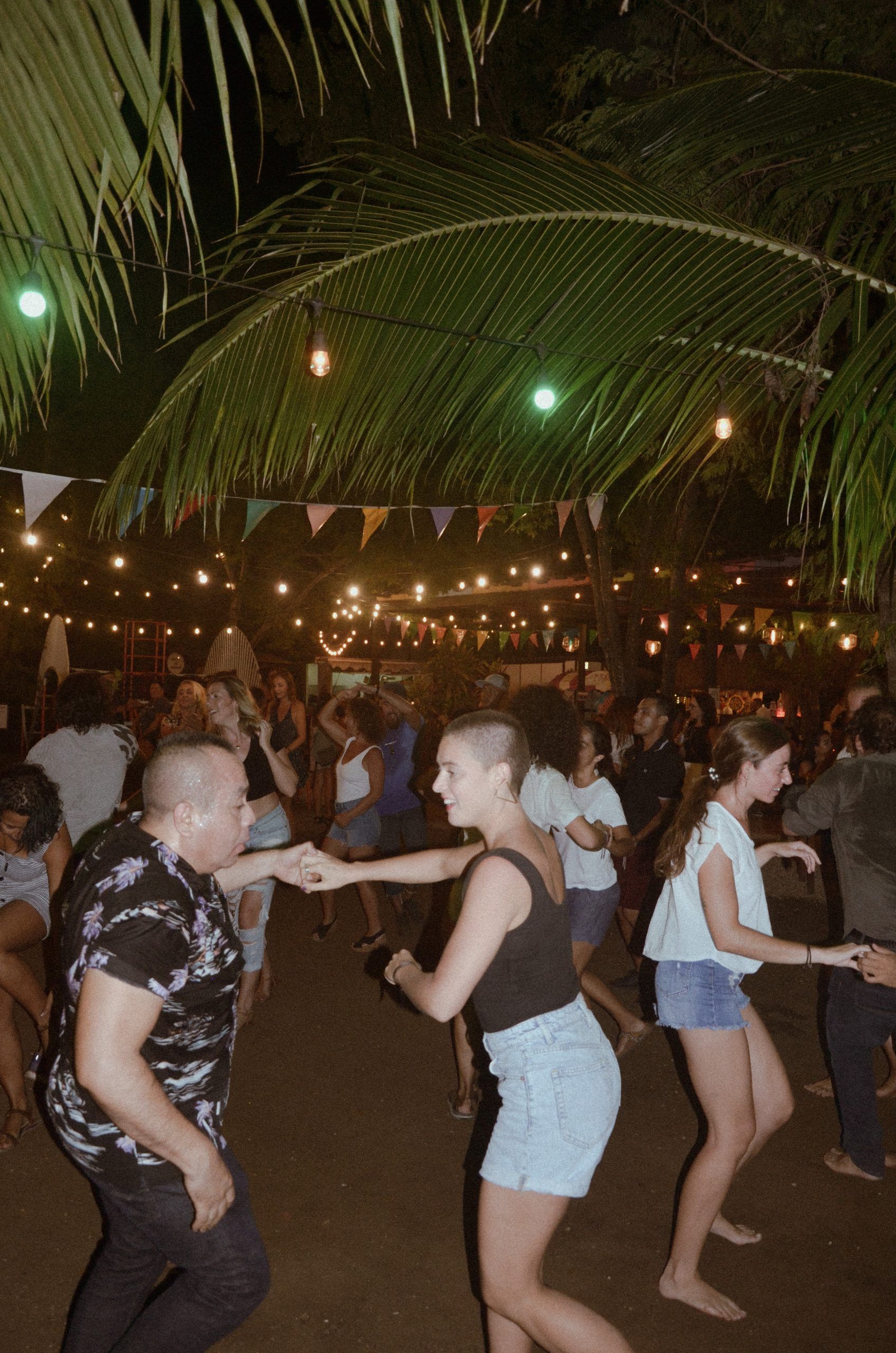 Gente bailando al aire libre por la noche, bajo luces de colores y palmeras.