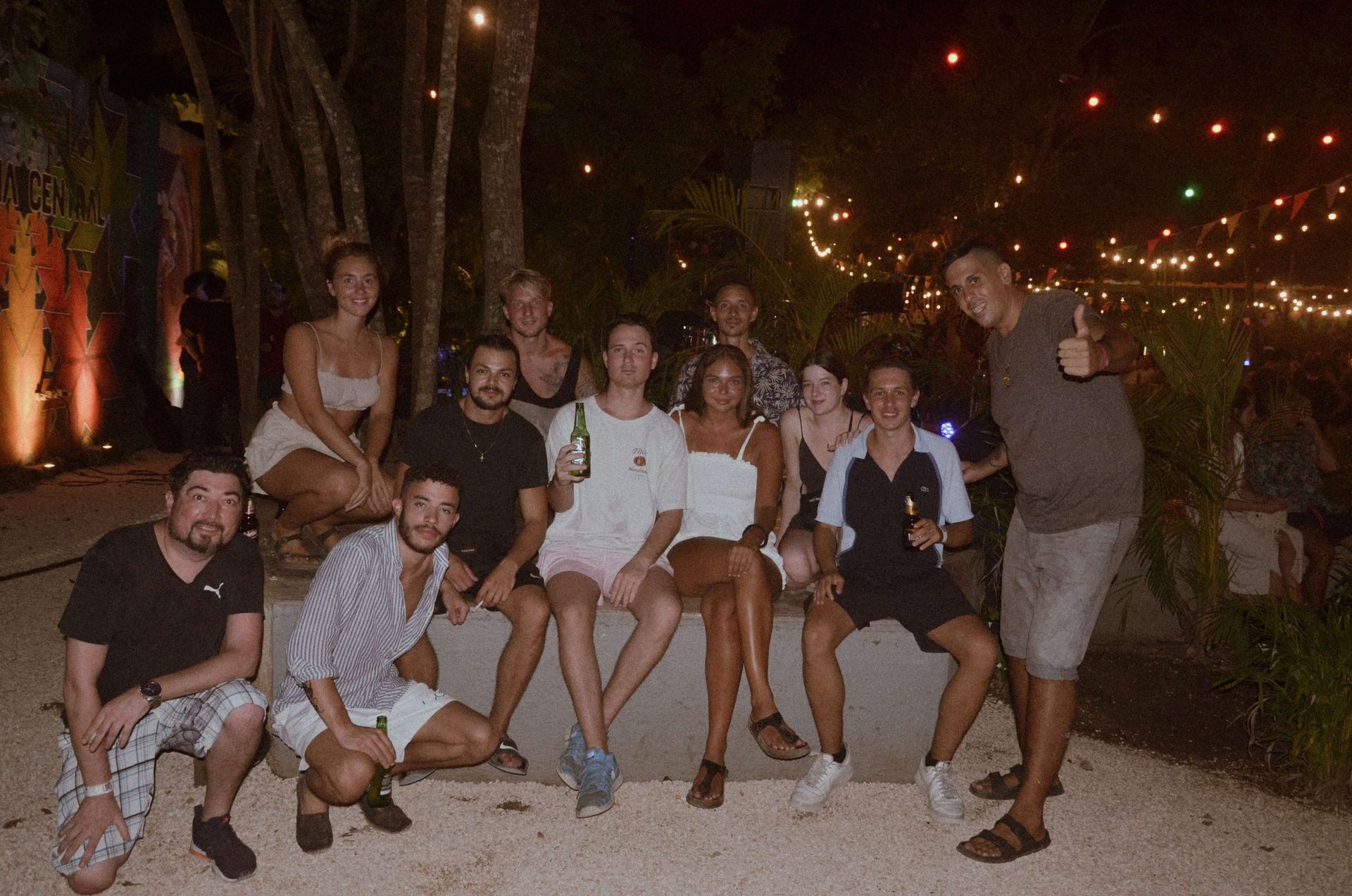 Grupo de personas sonriendo al aire libre por la noche, sentados en una pared con luces de cadena en el fondo.