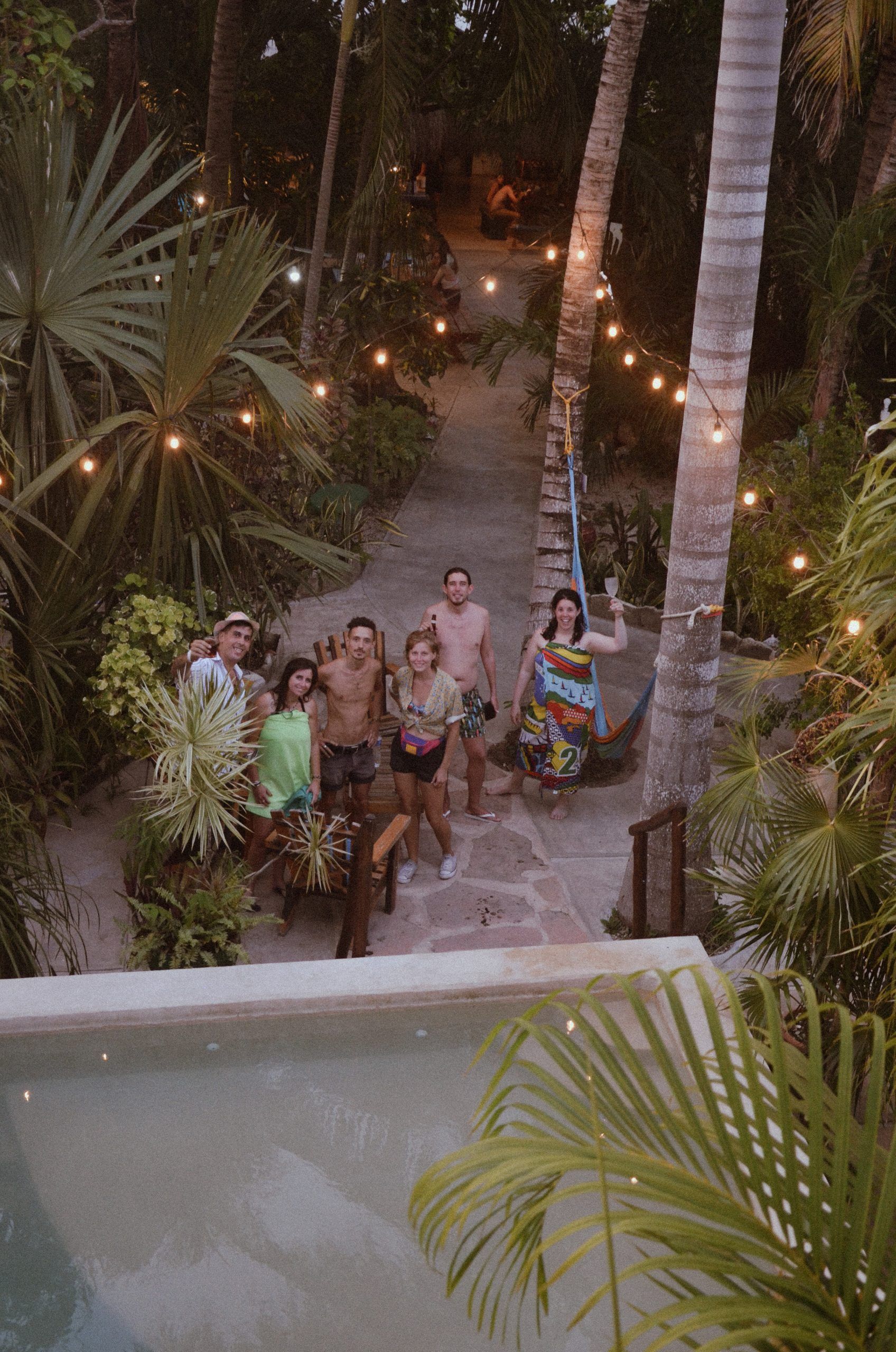 Personas posando en escalones de piedra junto a una piscina, rodeadas de exuberante follaje tropical y luces de cadena.