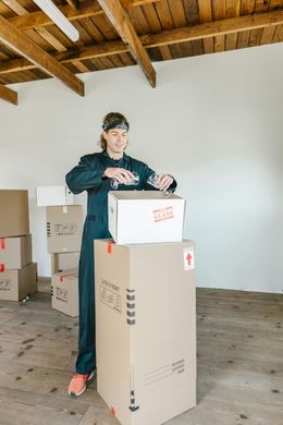 Person in blue jumpsuit taping a moving box, surrounded by other packed boxes, in a room with exposed wooden beams.