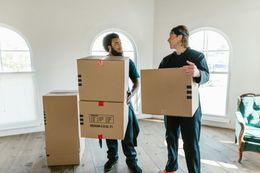 Two people in a bright room carrying cardboard boxes; preparing to move.