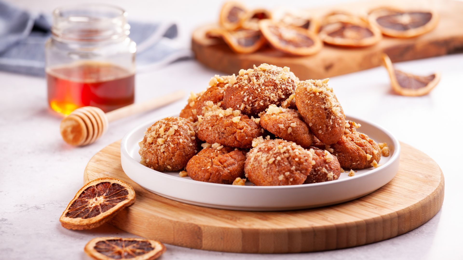 Plate of honey-soaked Greek cookies with chopped nuts, on a wooden board. Honey, dipper, and dried orange slices are nearby.