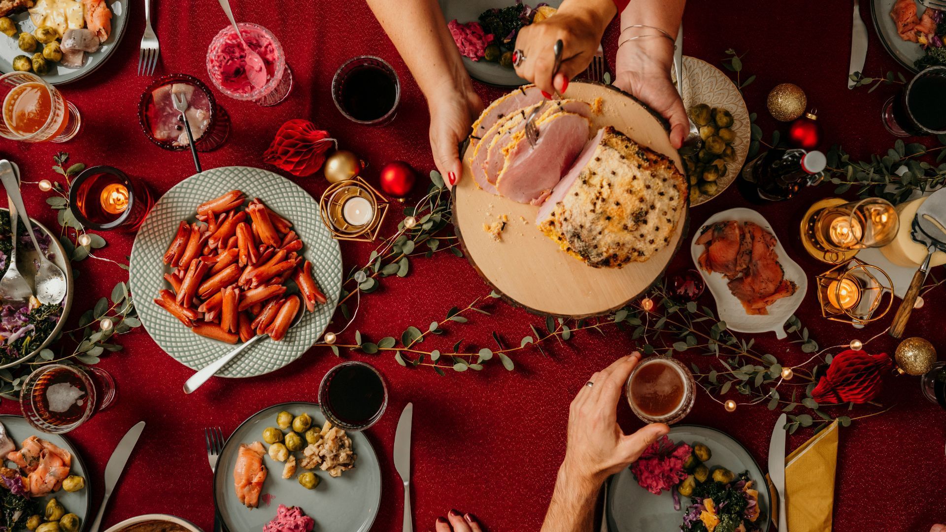 Holiday dinner table laden with food: pasta, ham, olives, drinks, candles, and hands serving.