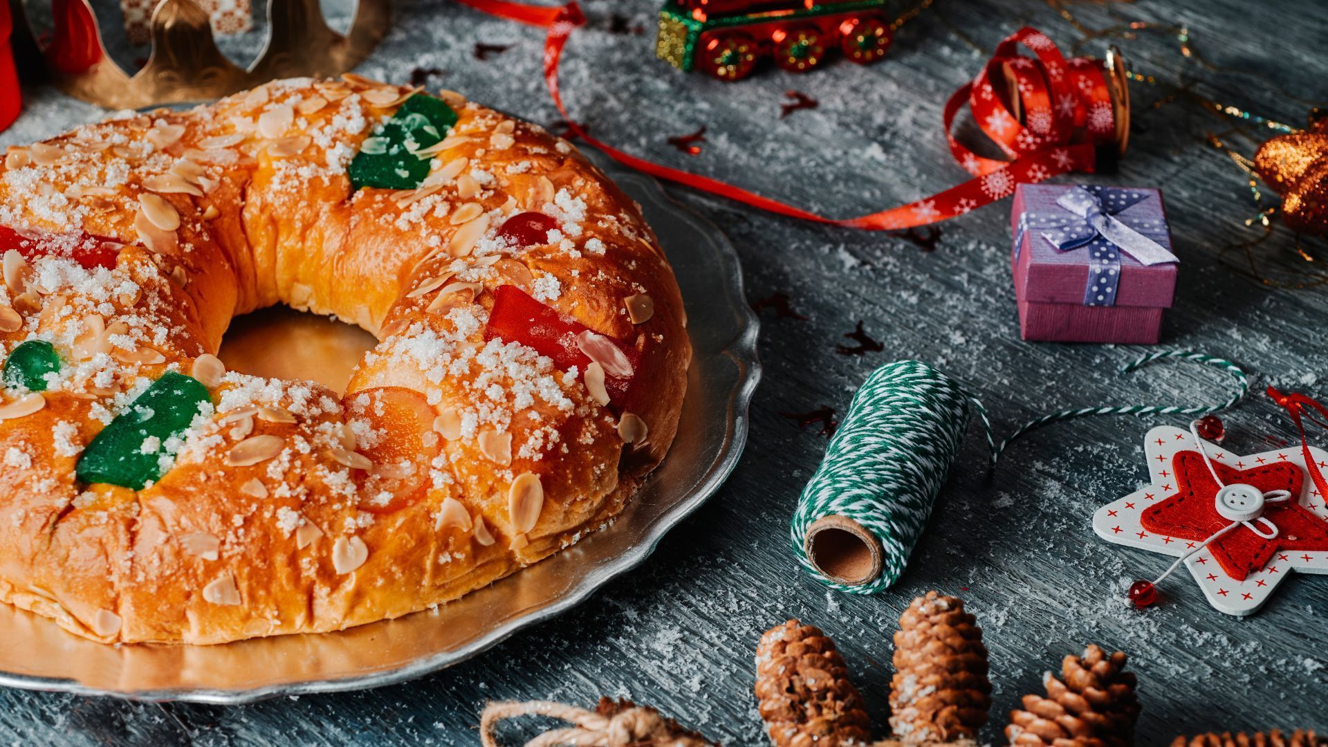 Ring-shaped pastry with almonds and candied fruit on a platter, surrounded by holiday decorations.