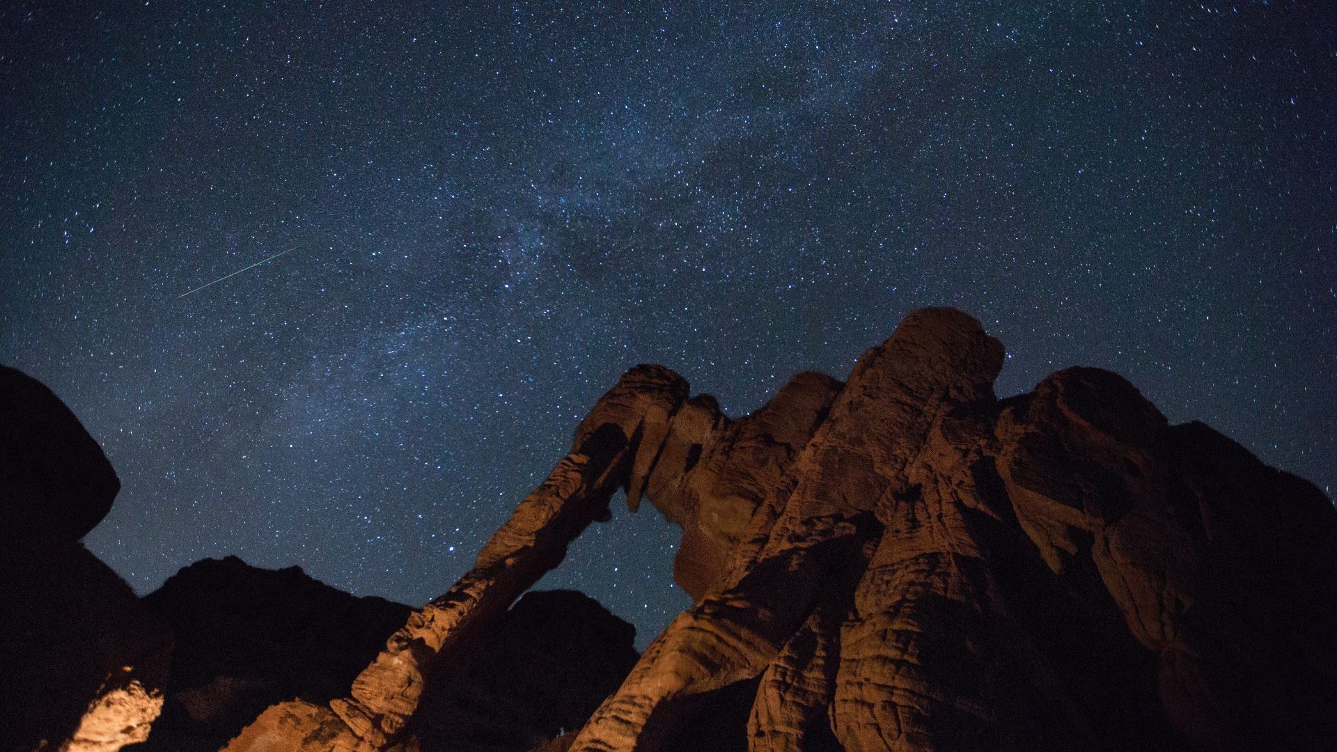 Night sky filled with stars above a sandstone arch in a desert landscape.