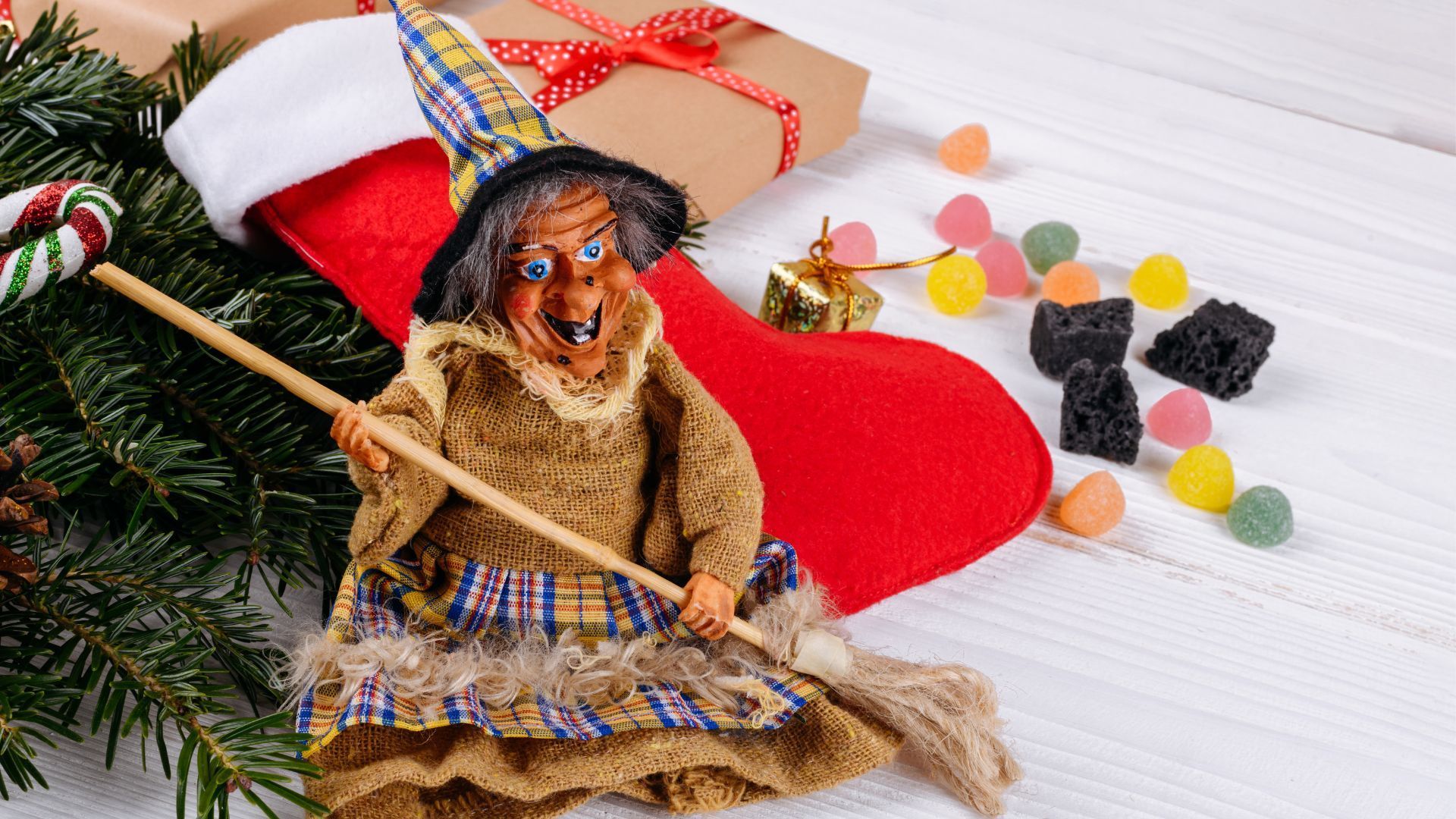 Befana doll on a broom near a stocking, gifts, and candy.