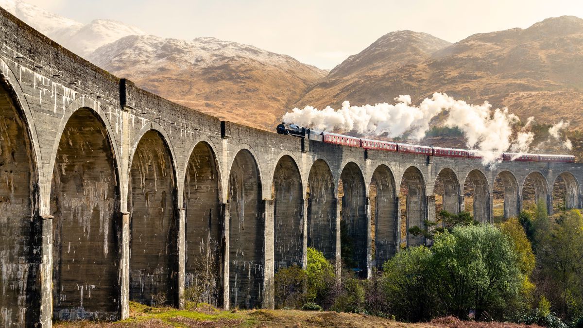 A train is going over a stone bridge with mountains in the background.