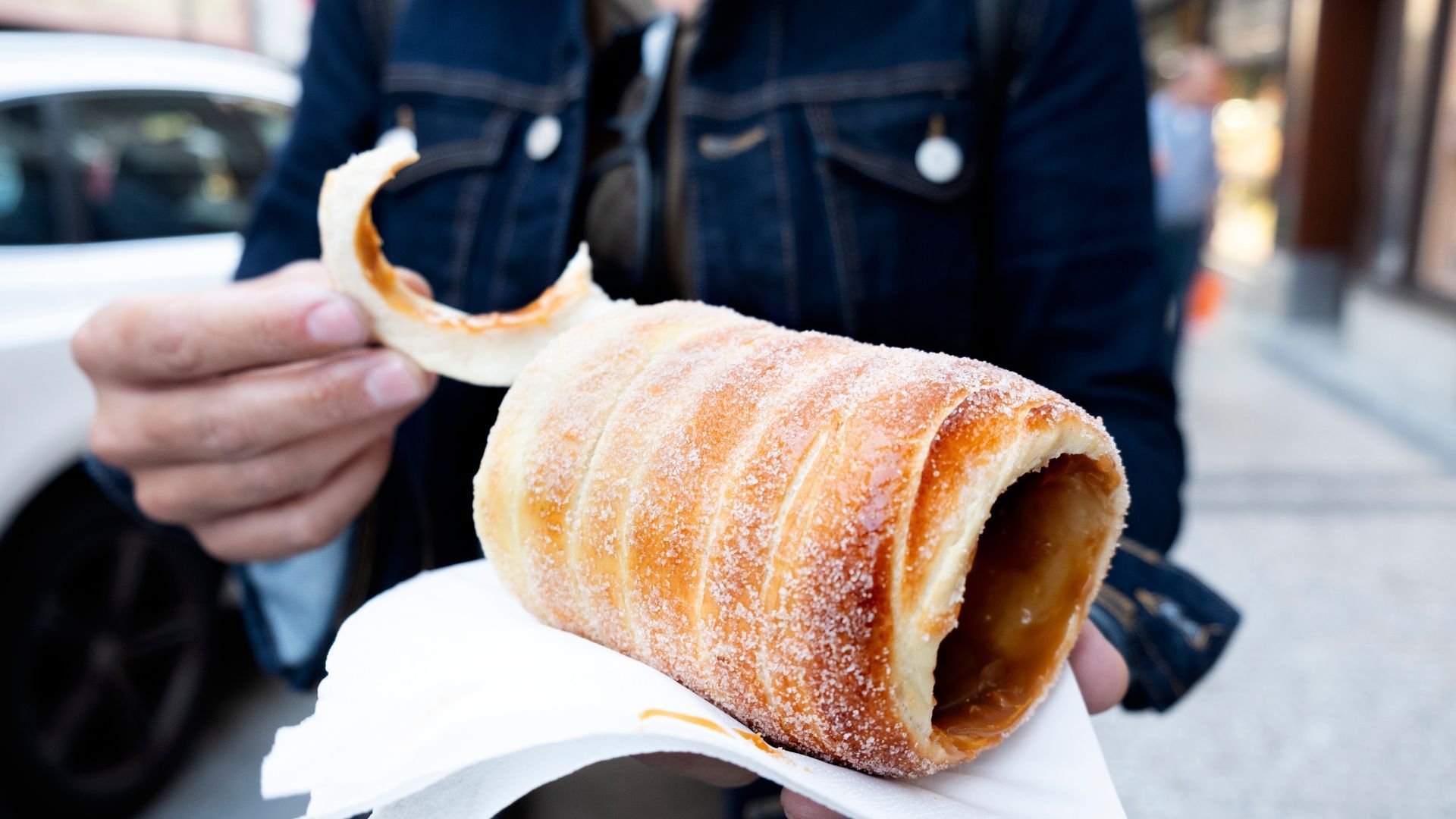 Person holding a Trdelník pastry, cylindrical, coated in sugar, partially eaten.
