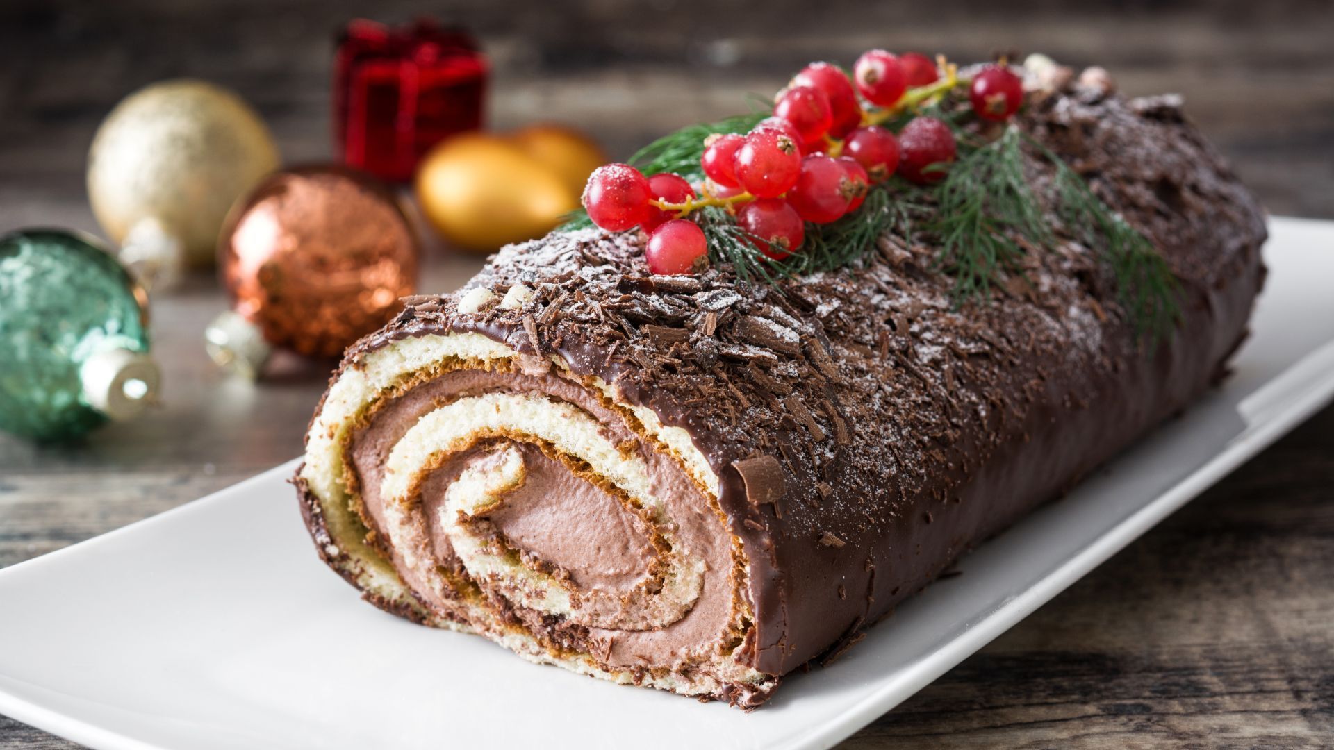 Chocolate Yule log cake with cream filling, topped with berries and sprigs on a white platter.