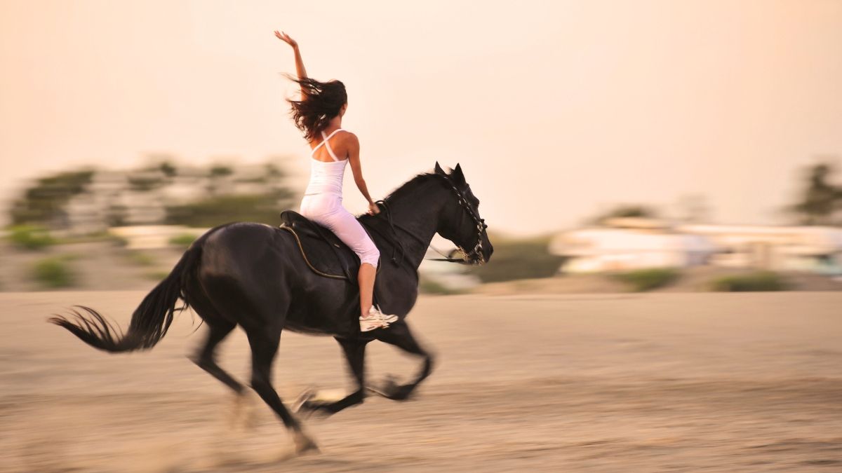 A woman in a white dress is riding a black horse on the beach.