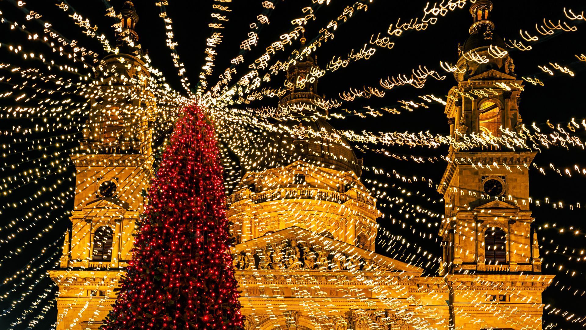 Christmas tree in front of a brightly lit building and lights against a black sky.