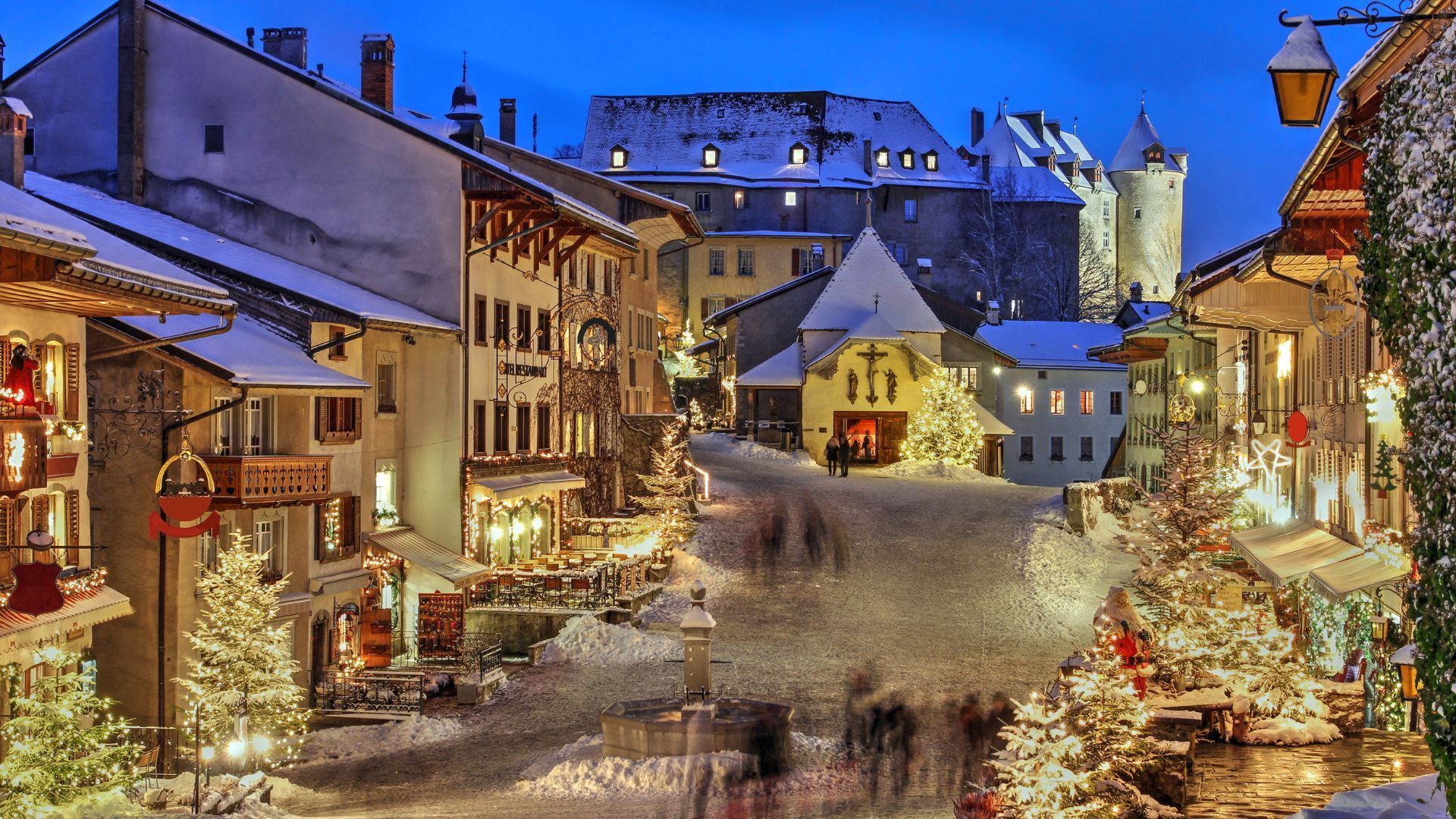 Snowy European village street at dusk, decorated for Christmas. Buildings, trees, and people are illuminated.