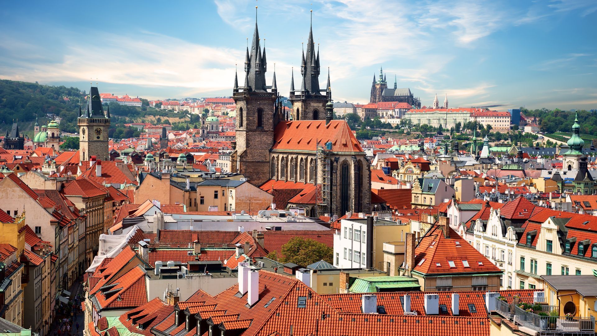 Prague cityscape with red rooftops, historic buildings, and the Church of Our Lady Before Týn under a bright blue sky.