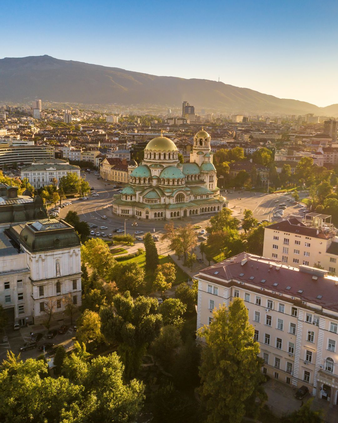 Sunlit cityscape with a turquoise-domed cathedral, trees, buildings, and mountains in the background