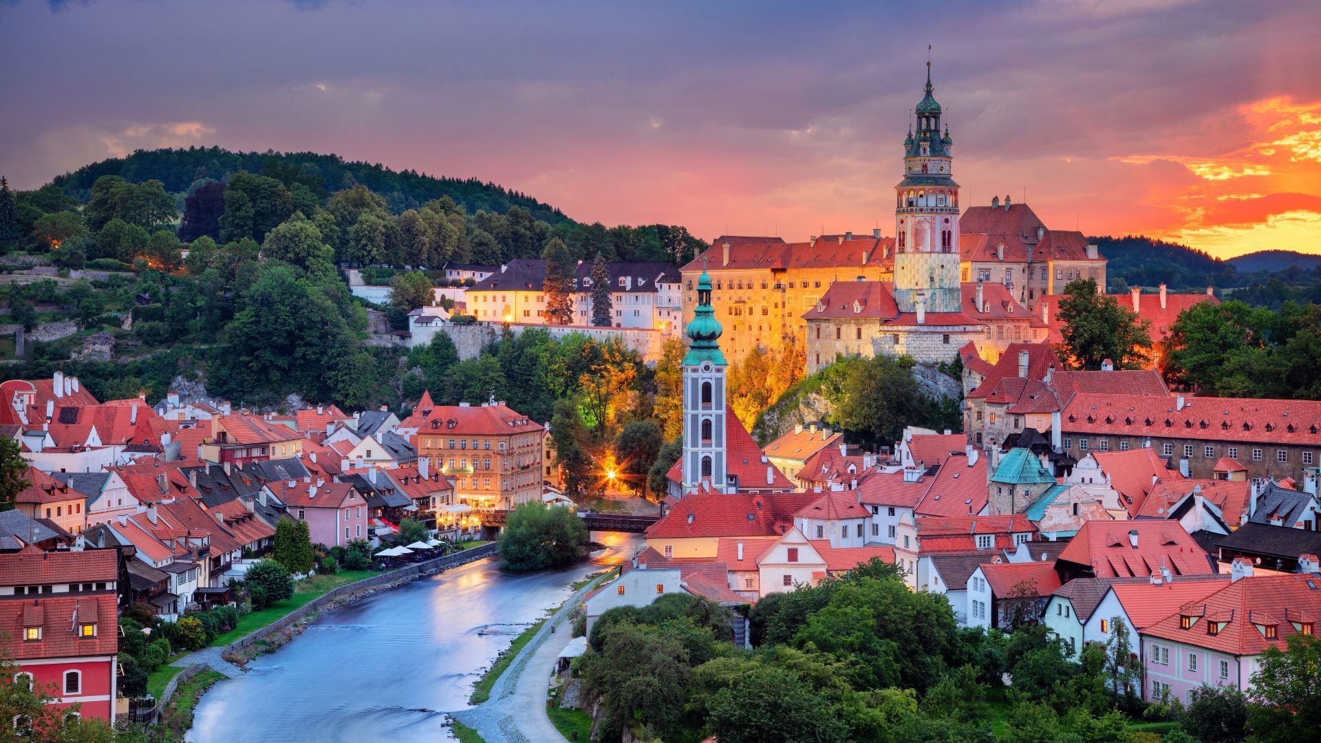Scenic view of Cesky Krumlov, Czech Republic, with red-roofed buildings, a river, and a tall church tower at sunset.