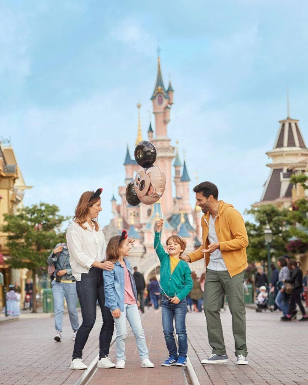 Family posing in front of a castle, with a child holding Mickey balloons at a theme park.