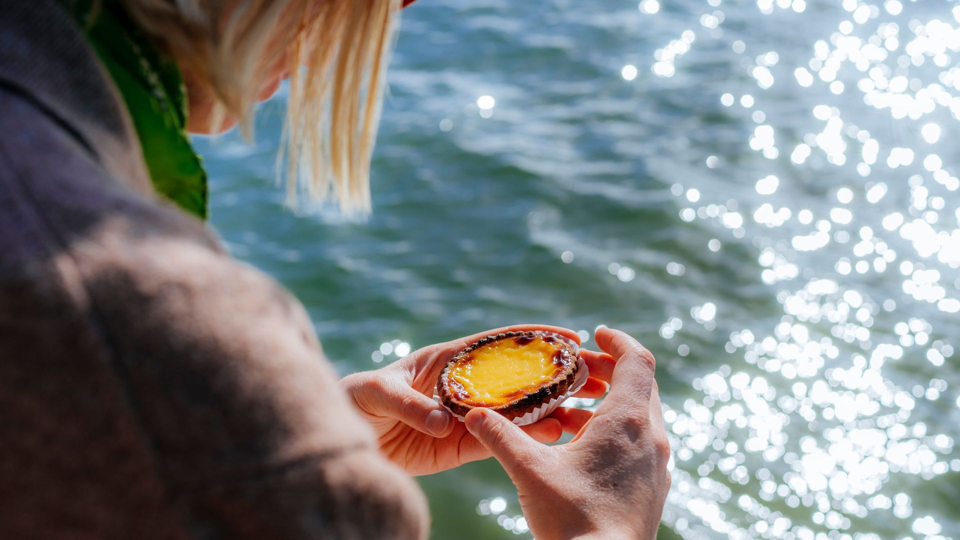 A person is holding a pastel de nata in front of a body of water.