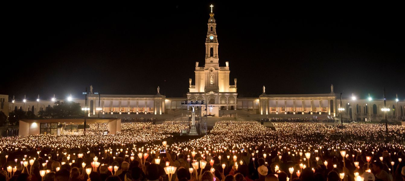 A large group of people are holding candles in front of a Fatima church at night.