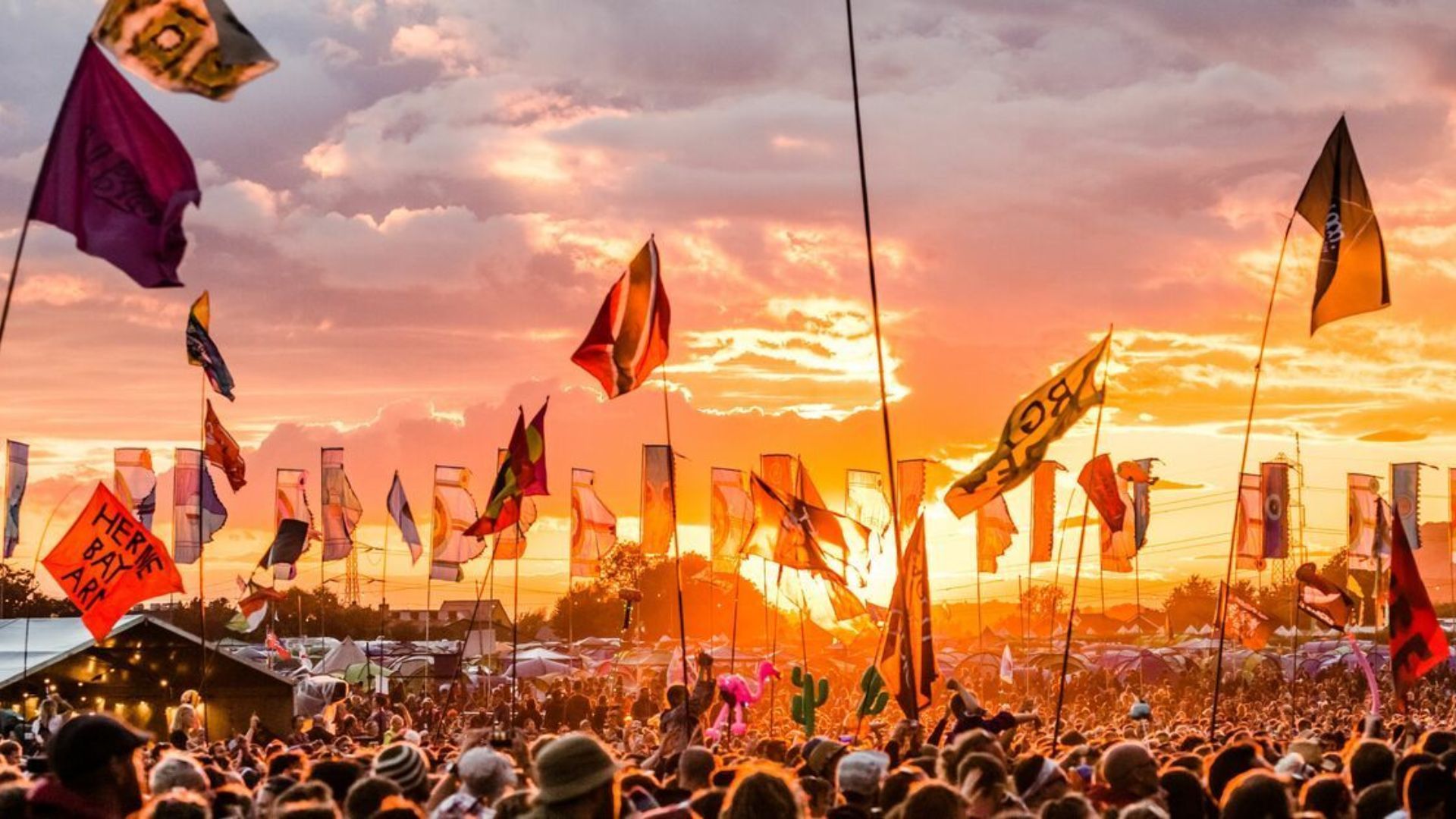 A vibrant music festival, Glastonbury in the UK crowd under a glowing sunset sky, filled with numerous colorful flags waving above.