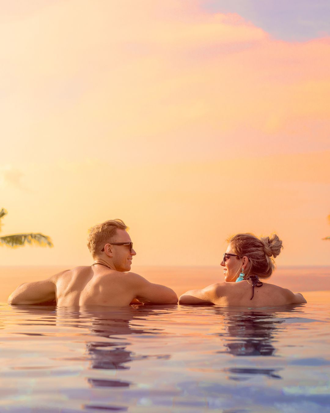 Two people relaxing in a pool at sunset, facing each other with warm orange sky reflected on the water