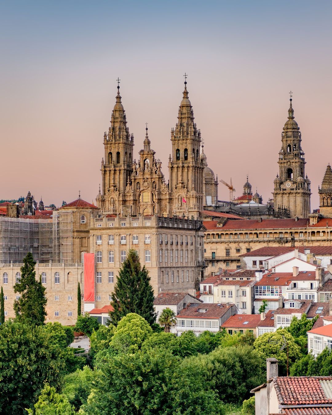Cathedral in Santiago de Compostela towers above a historic cityscape with red-tiled roofs and green trees at sunset.