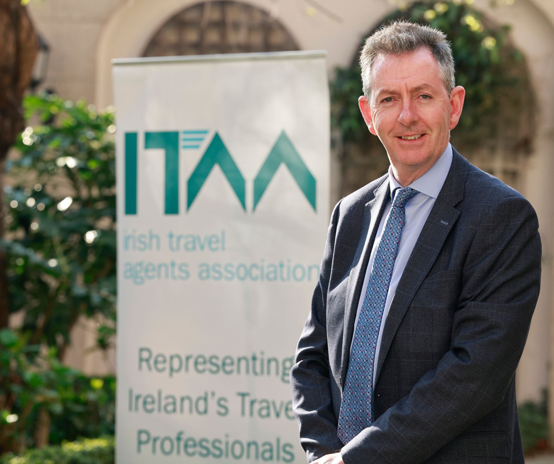 Tom Randles in a suit and tie stands in front of a sign that says irish travel agents association