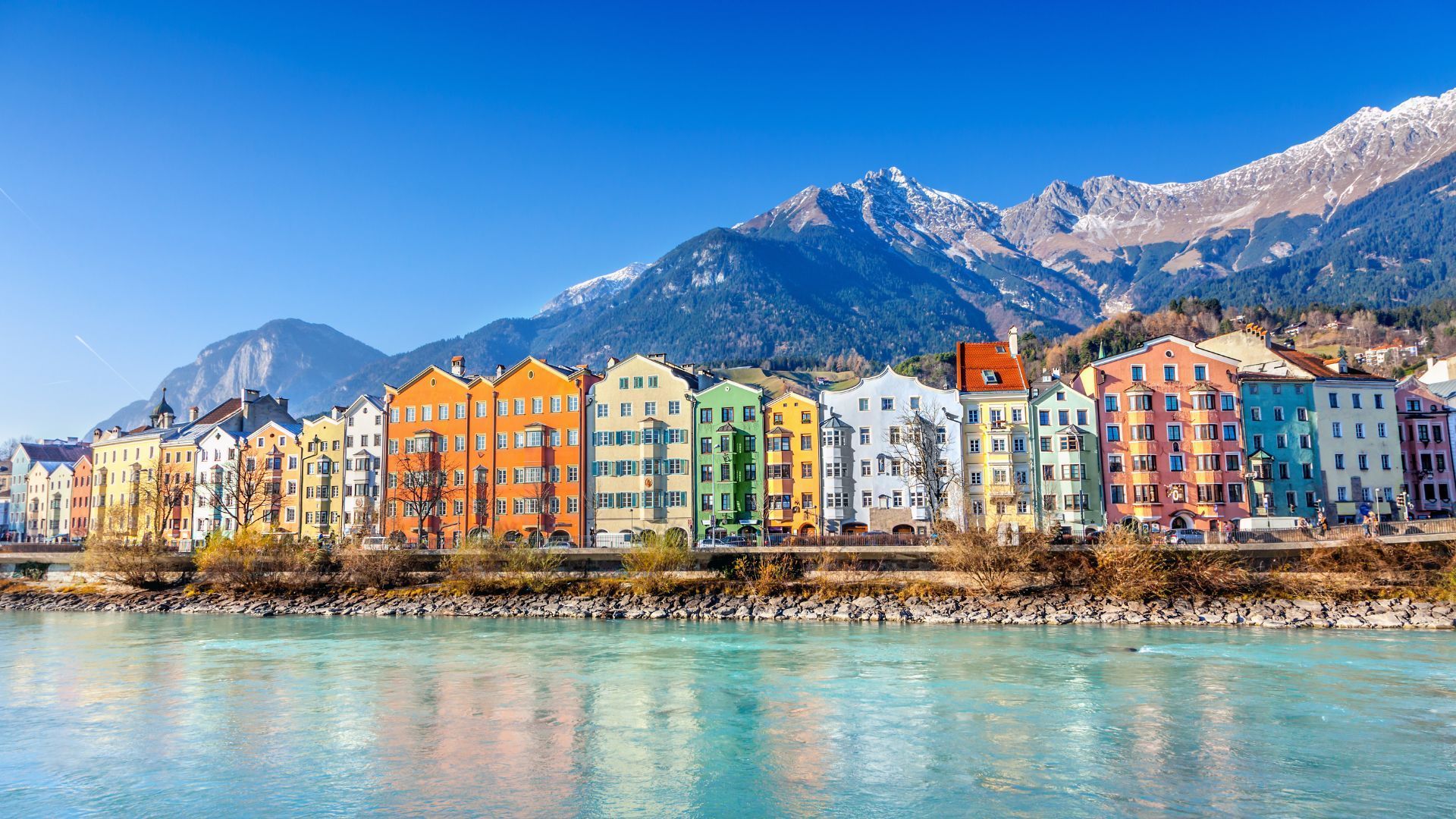 A row of colorful buildings in Austria along a river with mountains in the background.